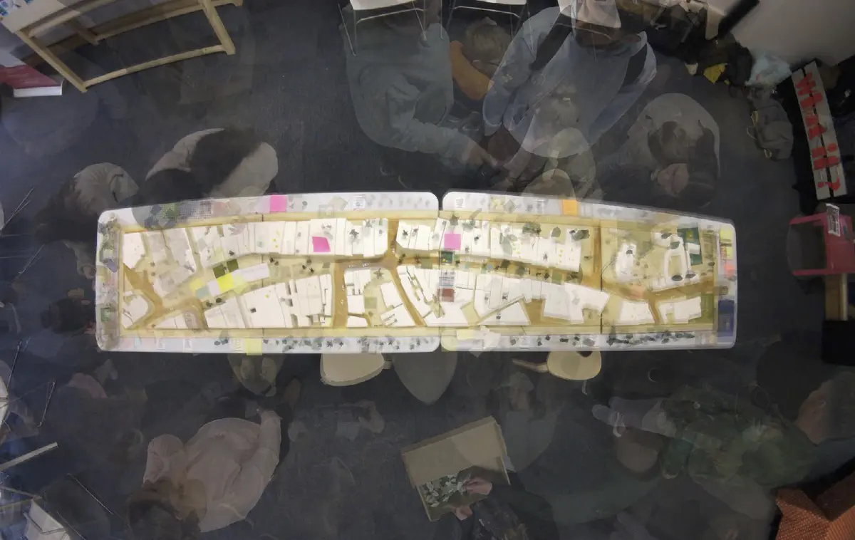 Looking to be the centrepiece of our exhibition, we proposed to create a site model of Dudley Highstreet at 1:200 scale with buildings modelled in a simple form. Local residents and visitors to the exhibition are then invited to decorate a chosen building to how they would envision it sat within the context of a greener Highstreet. We want to tie in our themes of biodiversity and materials by proposing that buildings can, and do, have the potential to be a lot healthier, sustainable, and interactive on the street. This site model also helps gain vital public opinions on the Highstreet and prompts discussion of areas most in need of attention.

To ensure that the model is utilising the MDF to the most efficient standard possible. First, the centres of the massing blocks have been hollowed out. This allows a reduction in materials used as other buildings can be cut in the centres of the model. Then the top is finished with a solid piece.
The waste material is placed into the recycling rack where other students can reuse it; otherwise, if the material left cannot be used for another project, it is recycled correctly by the university.

The model spanned just over 3.4m long and over 50cm wide. With the majority being cut from scrap woods that would fit the jigsaw of the shape, we were able to drastically cut down on waste. We also got some small benches and bee hives printed from recycled plastics, and used dried flowers as trees to represent the green spaces. Ultimately, the model seemed like a huge success, and really helped portray our ideas about Dudley High Street to those who came by. Everyone from Dudley who saw the model knew that it was the High Street, and interacted well with it. The size did make it difficult to move, especially to carry to the site, but it sat nicely in the centre of the the room and really drew people in to interact with it.

The model brought people in from outside to play with, write on, interact with. It became a spectacle for the public to come in and see what was happening and offer their ideas about what could be done.

The event was a success. Many people from the public (those who heard about the event, and those who just happened across it on the day) came in to interact with the model, which sat as the centre piece in the room. Kids liked to play with the pieces while the adults talked about things that could come from actions to help the High Street, not just leave it as is.

SOME OF THE KEY IDEAS WERE:
- The Shops need to be inviting for everyone. It’s no good having these places to go if people would rather take a bus to the next town.
- Just putting up boards across the windows is awful. Paint, Posters, Graffiti: Anything that actually makes the High Street look still alive is better than just wooden boards.
- More Green is needed. While it is obvious that there is a lack of Trees, many people wanted to push for these small bushes or planting spaces that could easily be implemented across the street.
- Spaces for public events or for people to at least gather at. While there is the Market and Fountain, it is not enough for the people of Dudley.
- Something needs to be done about the cars on the High Street. Some were Pro-Removal of all Cars, while some just wanted to limit when they could access the High Street.

Following the exhibition we created a series of drawings with our own interpretation of Sarah Wigglesworth ‘The Dining Table’ drawing. The drawings are a perfect representation of the our interactive model at the ERA Environmental Exhibition and Event. Showing the interaction the public had with the model, as well as the conversations that were held around the model. - Model - timelapse.png